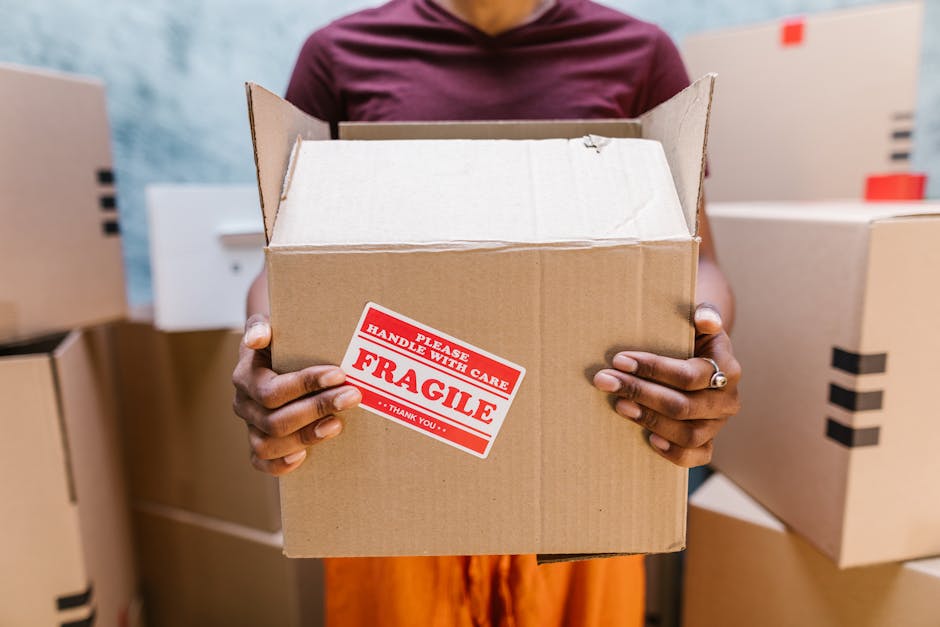 A person holding an open cardboard box labeled with a red and white 'Fragile' sticker that reads 'Please Handle with Care,' in a room filled with stacked moving boxes made of brown cardboard, some sea