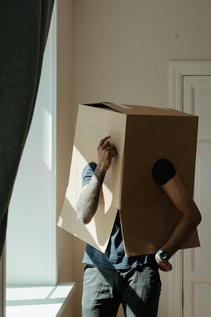 A person holding an open cardboard box labeled with a red and white 'Fragile' sticker that reads 'Please Handle with Care,' in a room filled with stacked moving boxes made of brown cardboard, some sea