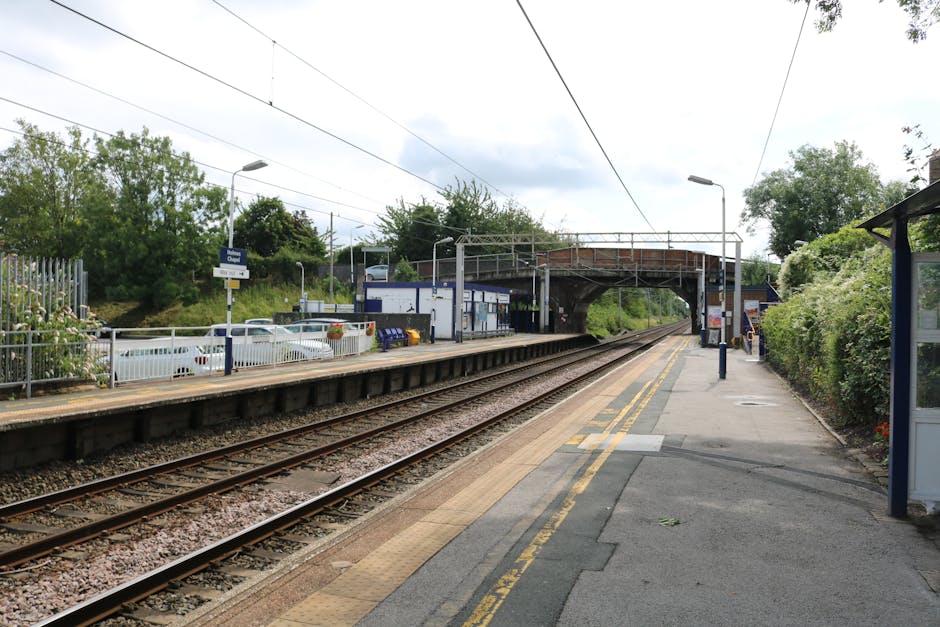 The image shows a train station platform adjacent to a railway track, with a metal railing separating the platform from the track area. There are several cardboard boxes and packed items placed on the platform, likely prepared for a home relocation or moving process. A small folding trolley and moving blankets are visible near the boxes, suggesting the transportation of household furniture. Parking spaces with multiple parked cars are visible alongside the station, behind the metal fence. Overhead electric wires run parallel to the tracks, supported by metal poles. A pedestrian overpass bridge extends over the railway, providing access between platforms, with a brick façade and metal railings. The scene is set during daytime with natural light, and lush green trees and bushes surround the station environment. A bus stop shelter is located on the right side of the platform, indicating public transport accessibility. The station is operated by Removal Companies Catford, supporting efficient house removals and moving logistics in the area, with the station's location and details relevant to local moving services and parking tips for the timing of relocation activities near Catford Bridge Station.