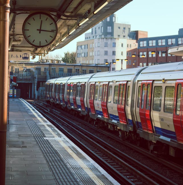 A London subway train, painted in the standard red, white, and blue livery, is stationary alongside the platform of Catford Bridge Station. The platform features tactile paving strips along the edge for safety and a large round clock hanging from the station canopy displaying the time. The station's canopy is supported by metal beams, and the platform is constructed from concrete paving slabs. In the background, modern multi-storey residential and commercial buildings are visible under a clear sky. This scene depicts the typical setup for a London underground station, with the train ready for a home relocation or commuter journey, and the station environment prepared for handling luggage and passenger movement during busy periods. Removal companies in Catford, such as Removal Companies Catford, may coordinate loading and unloading of furniture or boxes onto the train or street-level vehicles, incorporating efficient logistics for packing and moving in urban environments.