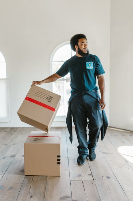 A man with dark skin, curly black hair, and a beard is standing inside a bright, empty room with natural light coming through a large arched window in the background. He is wearing a navy blue T-shirt with a logo on the chest, dark pants, and has a jacket tied around his waist. The man is holding a cardboard box with red tape on the sides, about to place it on a second, larger cardboard box that is already on the wooden floor. The larger box has a barcode label and black tape securing its edges, indicating it is part of a home relocation or furniture transport process. This scene depicts the packing and moving stage of a house removal, and the setting is typical of a professional removals company such as Removal Companies Catford, which specializes in efficient furniture transport and packing for tight access locations in areas like Catford. The room appears to be an unfurnished, minimally decorated space suitable for staging moving activities.
