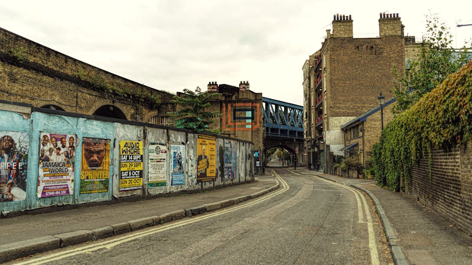 A narrow street scene in an urban residential area with a gently curving asphalt road featuring double yellow lines on both sides, indicating no parking zone. On the left, a long weathered metal fence covered with colorful posters and advertisements for local events and concerts runs alongside the sidewalk, which is lined with small concrete curbs. In the distance, a blue metal bridge passes over the street, supported by brick and stone structures, indicating an underpass or railway bridge. To the right, a brick wall topped with green foliage borders the sidewalk, leading to multi-storey brick buildings with chimneys and balconies seen further along the street. Overcast skies cast diffuse light over the scene, capturing a typical day of urban house removals or packing and moving activities, with Removal Companies Catford potentially involved in this area, as suggested by the context.