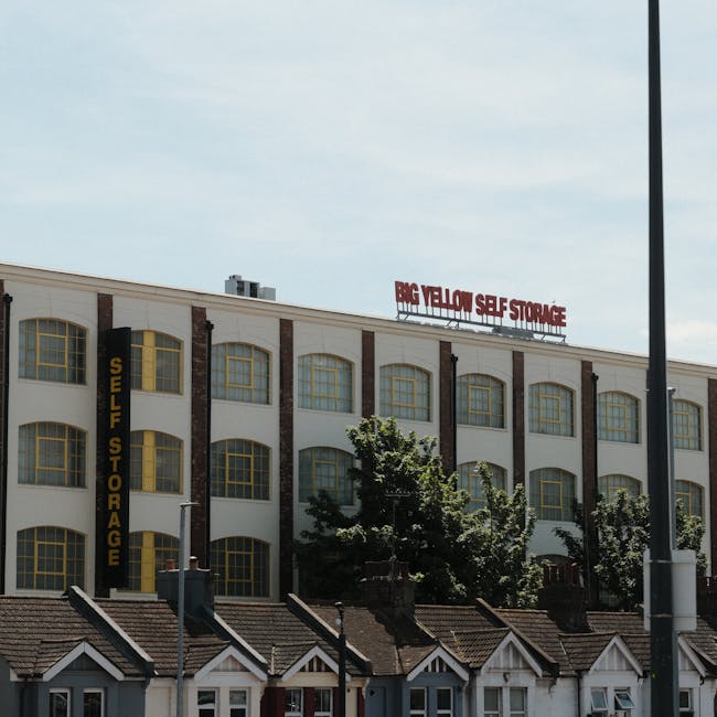 A three-storey commercial building painted white with brown brick accents, featuring large yellow-framed windows on each floor. A vertical black sign reading 'SELF STORAGE' is attached to the left side of the building. On the roof, a red sign displays 'BIG YELLOW SELF STORAGE.' In front of the building, there is a row of small residential-style houses with sloped roofs and chimneys, partially obscured by green trees. The sky above is partly cloudy with patches of blue. The scene appears to be in an urban or suburban area, relevant to house removals or storage services, illustrating a typical location where moving and storage logistics, such as loading or unloading, may take place, supporting content about home relocations and furniture transport through an external building setting.