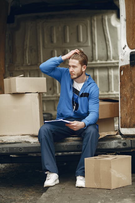 A young man with light skin and short, styled hair sits on the edge of an open van with a textured metal interior, surrounded by neatly packed cardboard boxes of various sizes, some on the ground and 